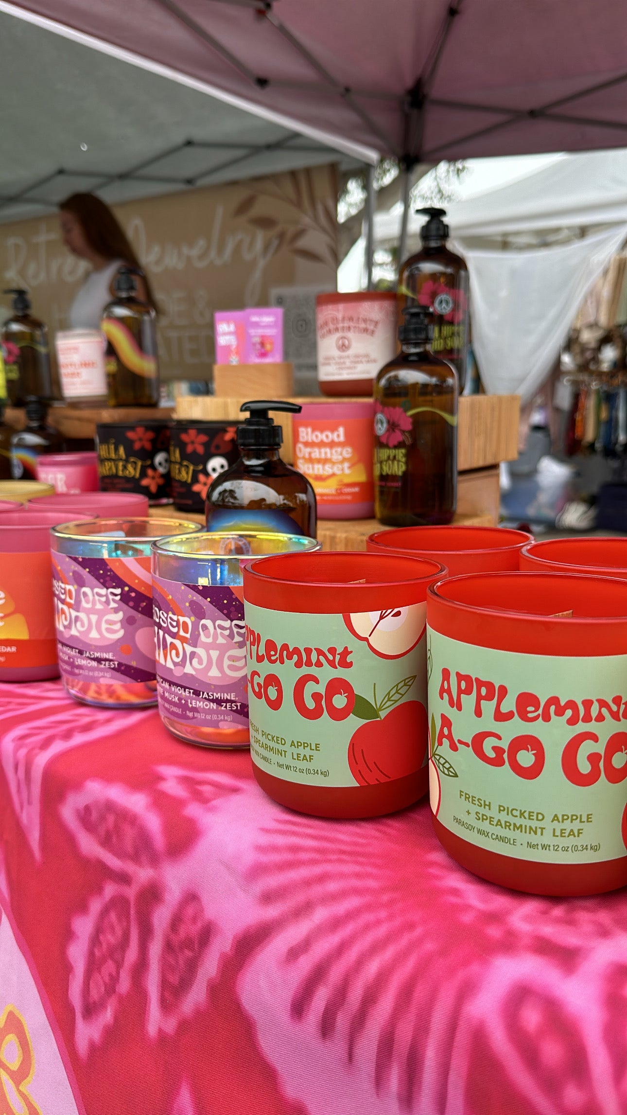 Applemint A-Go Go products on a farmer's market table with a pink tablecloth and various bottles in the background.