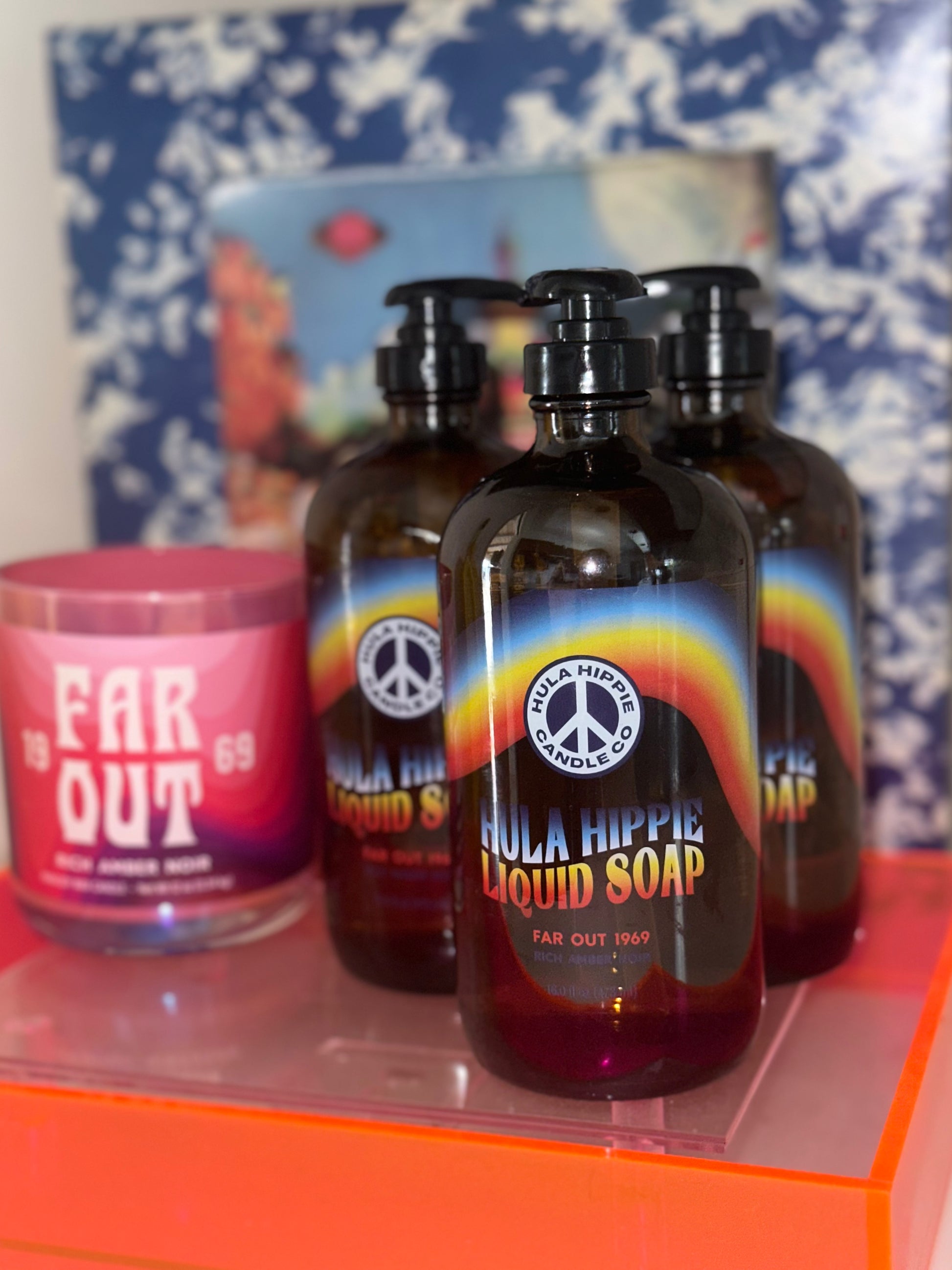 Three amber glass bottles of Far Out Liquid Soap with colorful labels, displayed on a surface with a floral pattern and a small container in the foreground.