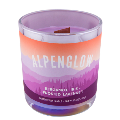 A scented candle labeled "Alpenglow" and scented notes of bergamot, iris, and frosted lavender, in a clear glass jar with a purple to orange gradient design.