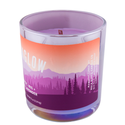 Right-side view of a scented candle labeled "Alpenglow" and scented notes of bergamot, iris, and frosted lavender, in a clear glass jar with a purple to orange gradient design and a forest scene.