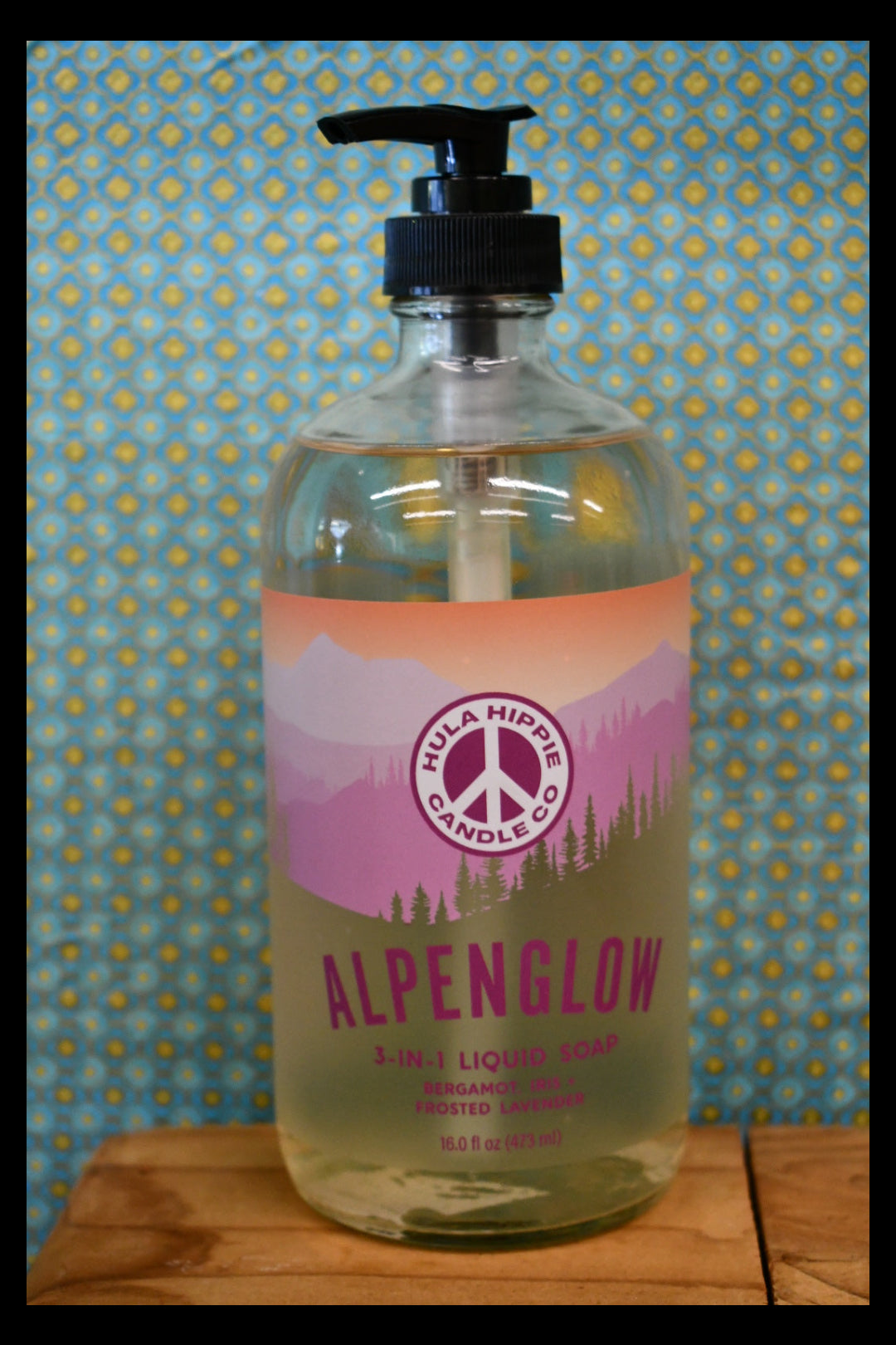 A bottle of Alpenglow 3 in one Liquid Cleanser by Hula Hippie Candle Co., lying on a blue and yellow patterned surface next to a wooden surface.