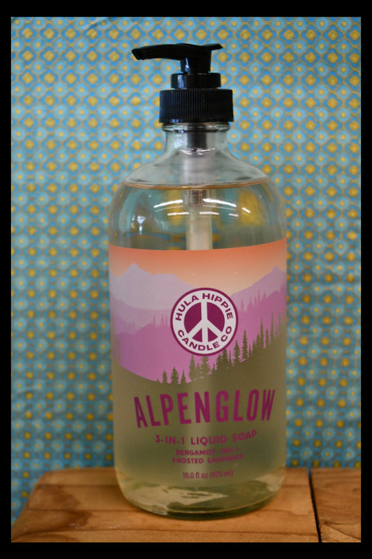 A bottle of Alpenglow 3 in one Liquid Cleanser by Hula Hippie Candle Co., lying on a blue and yellow patterned surface next to a wooden surface.