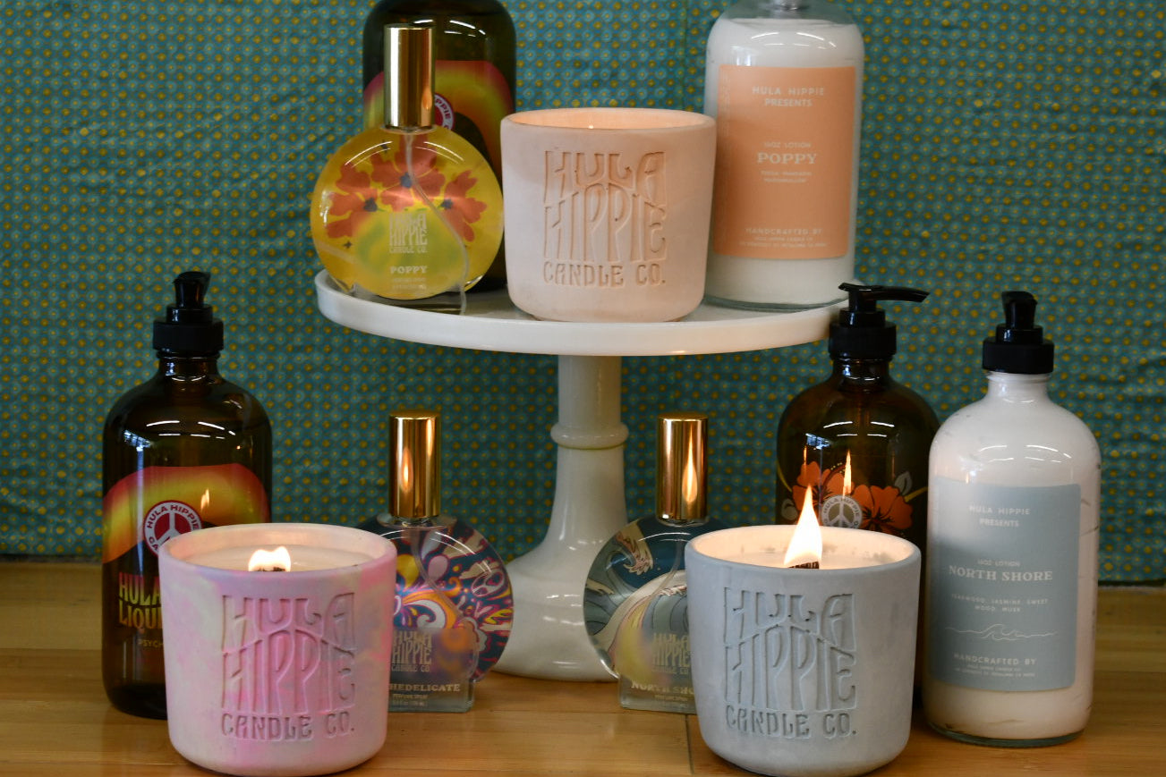A collection of bath and body products from hula Hippie Candle Co. arranged on a white two-tiered pedestal stand and tabletop against a green patterned backdrop.